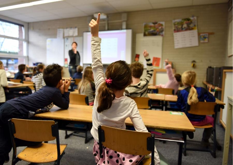 Children with teacher in a classroom. Some children with their hands up.