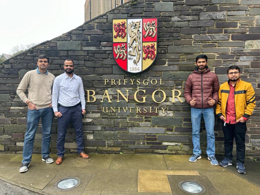 Four students standing in front of a wall with the Bangor University crest 