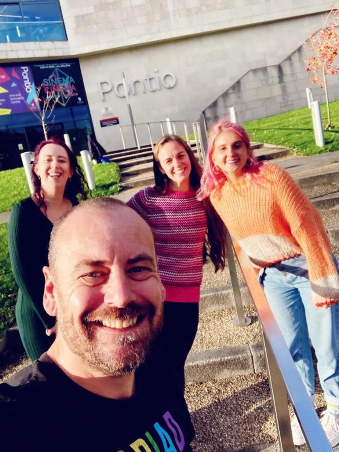 Image of 4 members of the Annedd Ni/Blas Pontio team outside Pontio building in Bangor. The image shows a man in a black t-shirt smiling, behind him are 3 women wearing green, pink and orange. In the background, you can see the steps leading up to Pontio and the Pontio sign