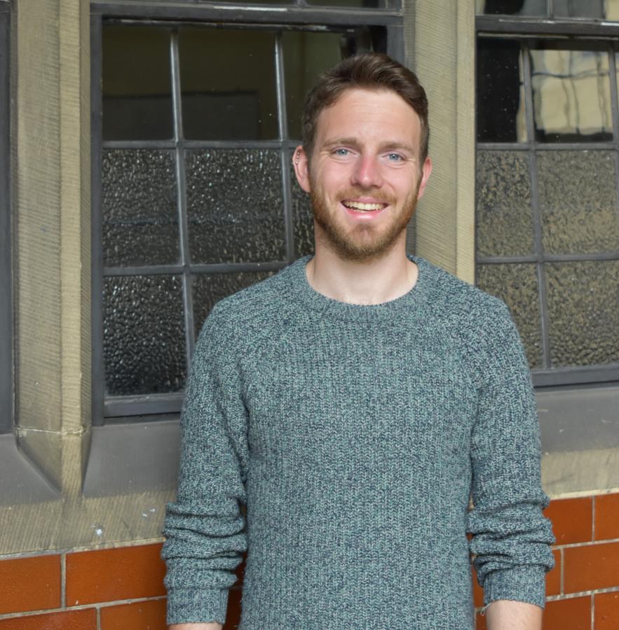 A picture of Alex Ioannou smiling in front of a window in the Main Arts Building of Bangor University.