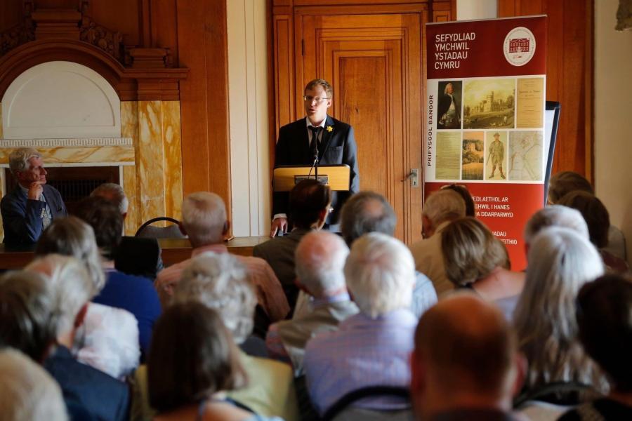 Dr Shaun Evans stands behind a lectern giving a talk to a room full of people, with an ISWE banner on display next to him. 