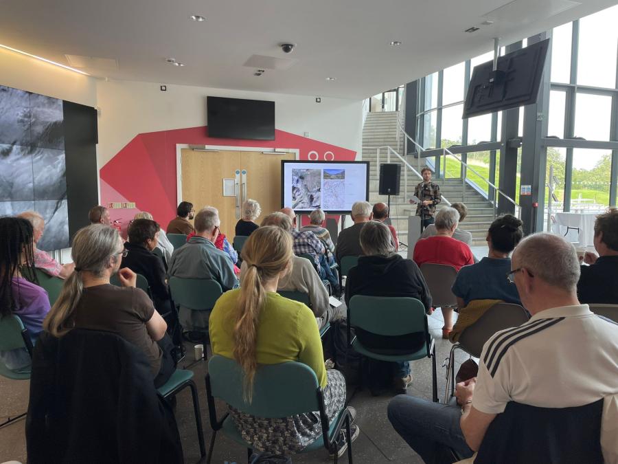 A photo of Alex Ioannou presenting to an audience, with a powerpoint presentation displayed on a large screen