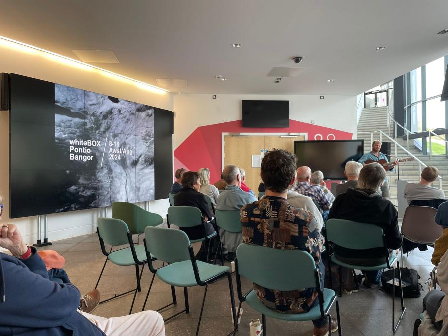 A photograph of musician Gwilym Bowen performing to a thoroughly engaged audience, with the landscape of Dyffryn Ogwen visible on a nearby screen