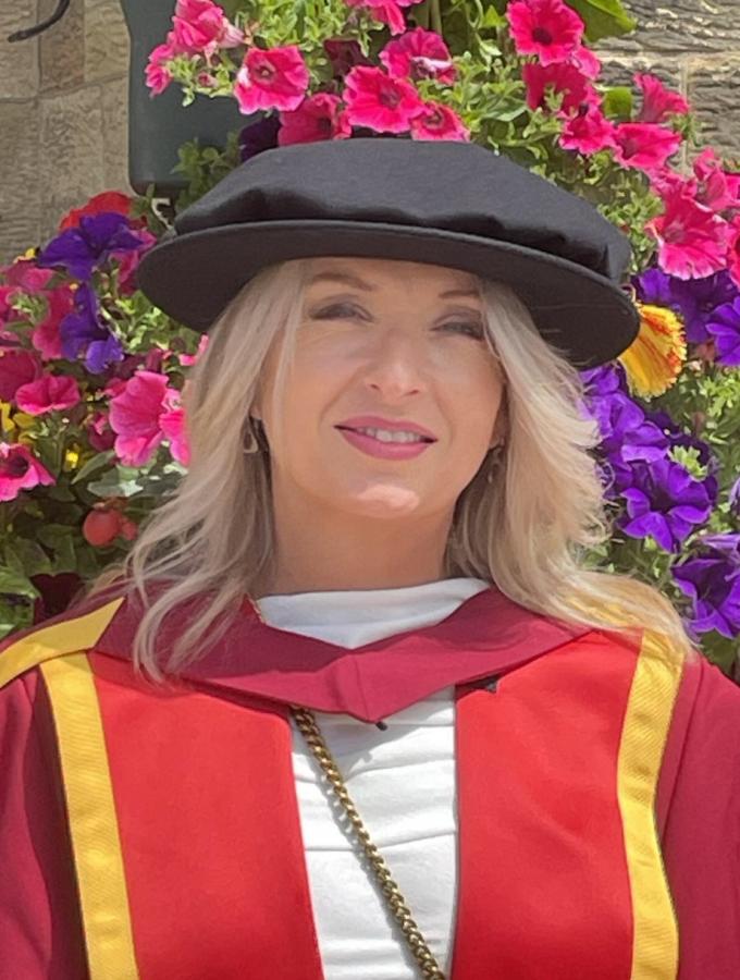 A photograph of Dr Meinir Moncrieffe, posing in her doctoral graduate robes during her graduation, in front of an array of colourful flowers