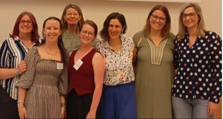 7 women stand in a row smiling. There is no visible background and all are wearing semi-formal conference clothes