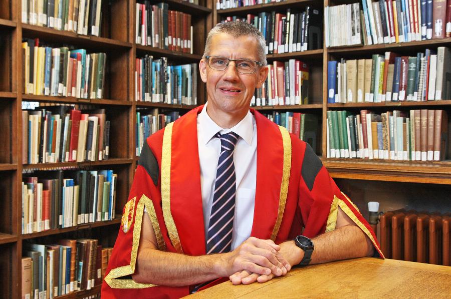 A man wearing glasses sitting down wearing red and gold graduation robe with bookshelves in background