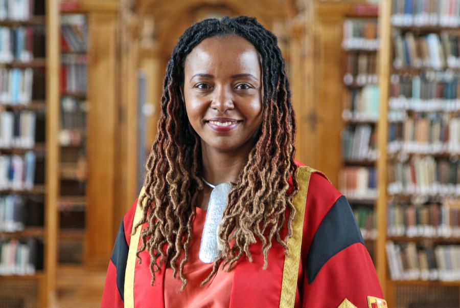 A woman wearing red and gold graduation robe standing in a library with bookshelves in the background.