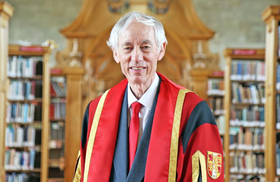 A man wearing wearing red and gold graduation robe with bookshelves in background