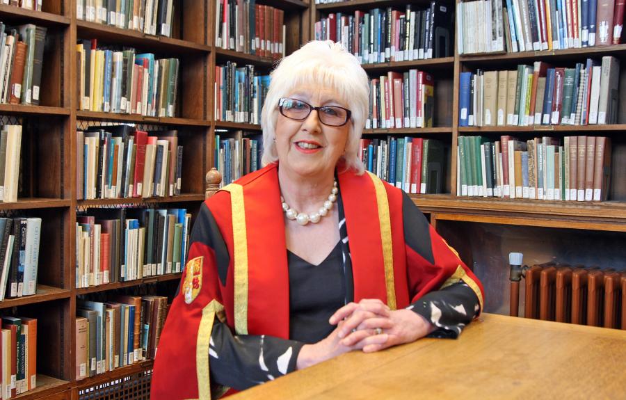 A female wearing glasses sitting down wearing red and gold graduation robe with bookshelves in background