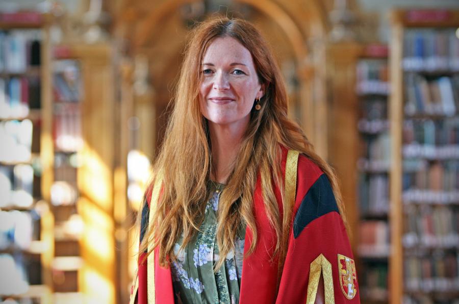 A female wearing wearing red and gold graduation robe with bookshelves in background