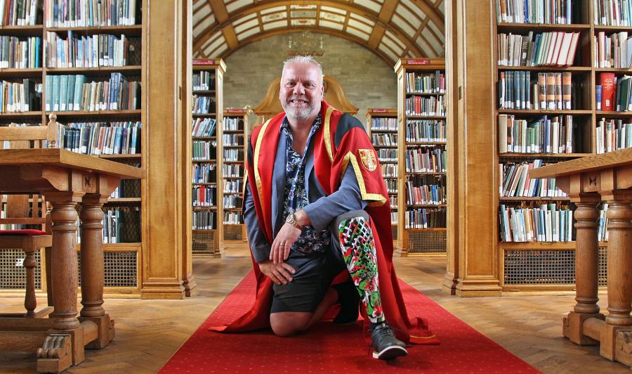 A man wearing red and gold graduation robe kneeling down. Right leg is prosthetic with a red and green Welsh design. He is in a library with bookshelves in the background.