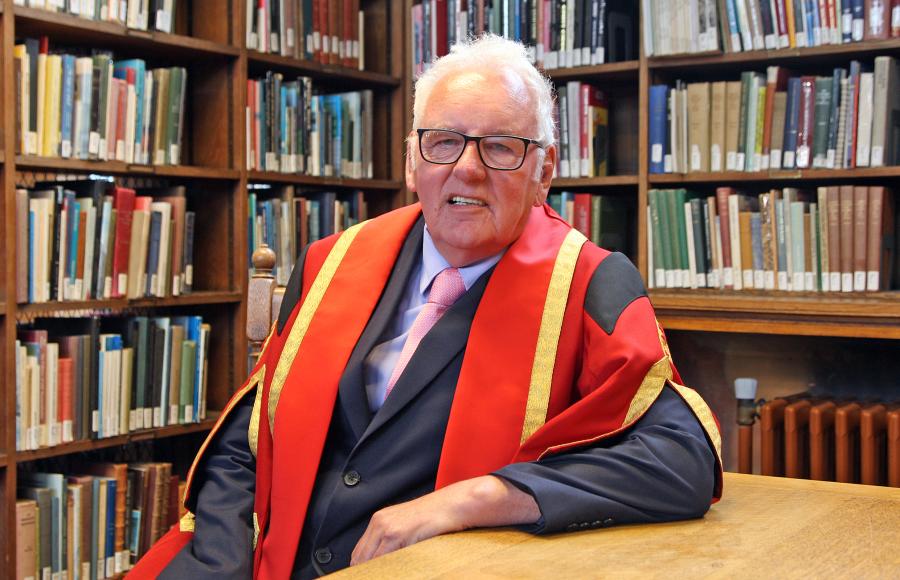 Image of a man wearing glasses and red and gold graduation robe. His right arm leans on the table before him. There are bookshelves in the background.