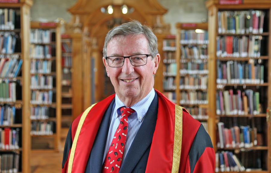 A man wearing glasses and red and gold graduation robe standing with bookshelves in the background