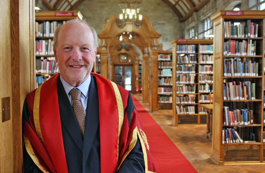 A man wearing red and gold graduation robe leaning on wooden pillar in a library with bookshelves in the background