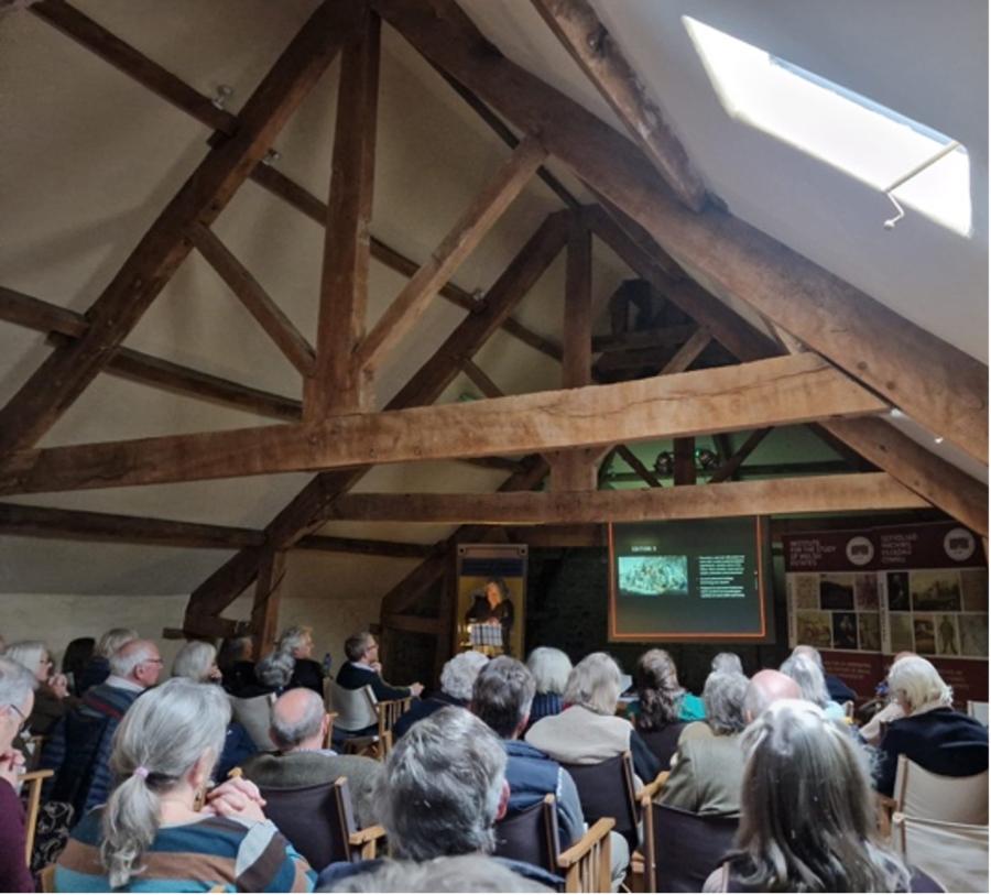 An attic room with an exposed wooden roof structure and an audience listening to a presentation. 