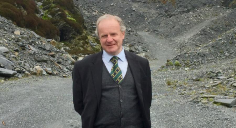 A photograph of David Gwyn smiling, behind him is an example of the slate landscape, with a path that is flanked by slate slag heaps 