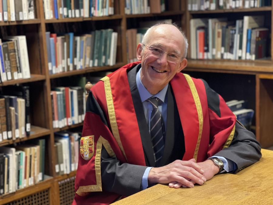 Image of Nigel Brown wearing red and gold honorary graduate robes sitting at a table